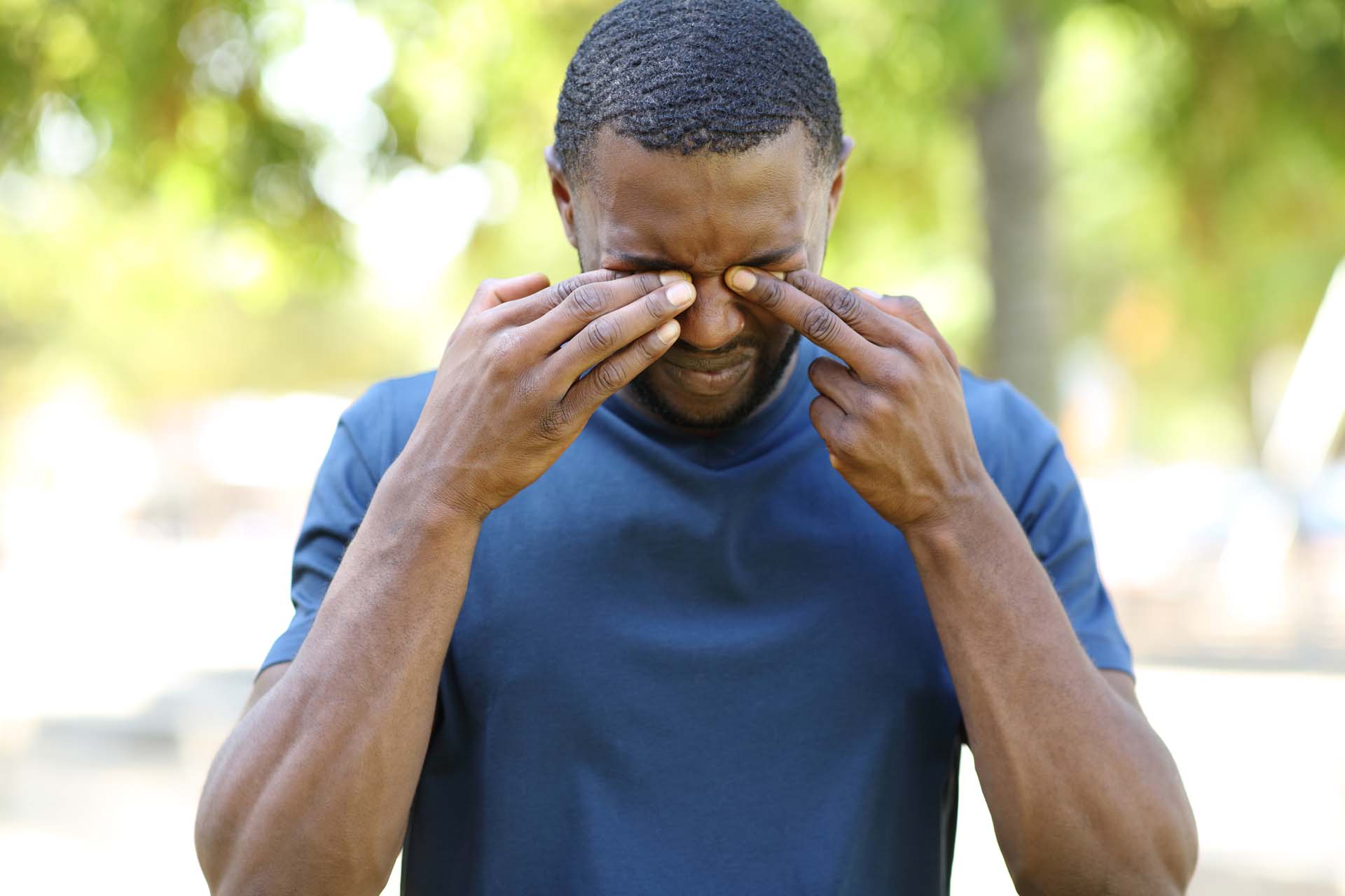 Front view portrait of a man with black skin scratching itchy eyes in a park